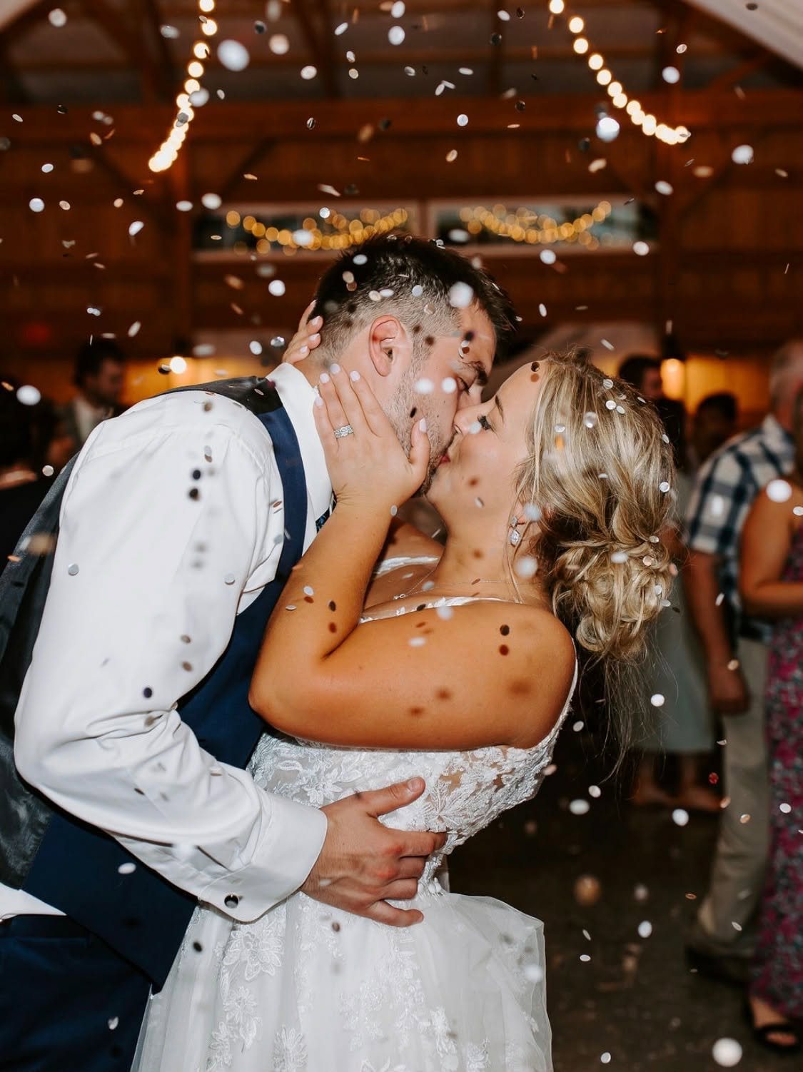 Newlyweds kissing under confetti and string lights in the barn at Sunset Meadows